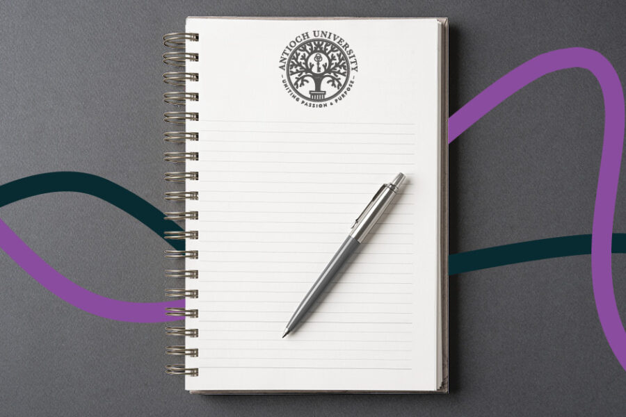 An open spiral notebook with lined pages and a silver pen resting on it. At the top of the page is the Antioch University logo with the words “Uniting Passion & Purpose.” The notebook is set on a gray background with purple and teal wave-like lines flowing across it.