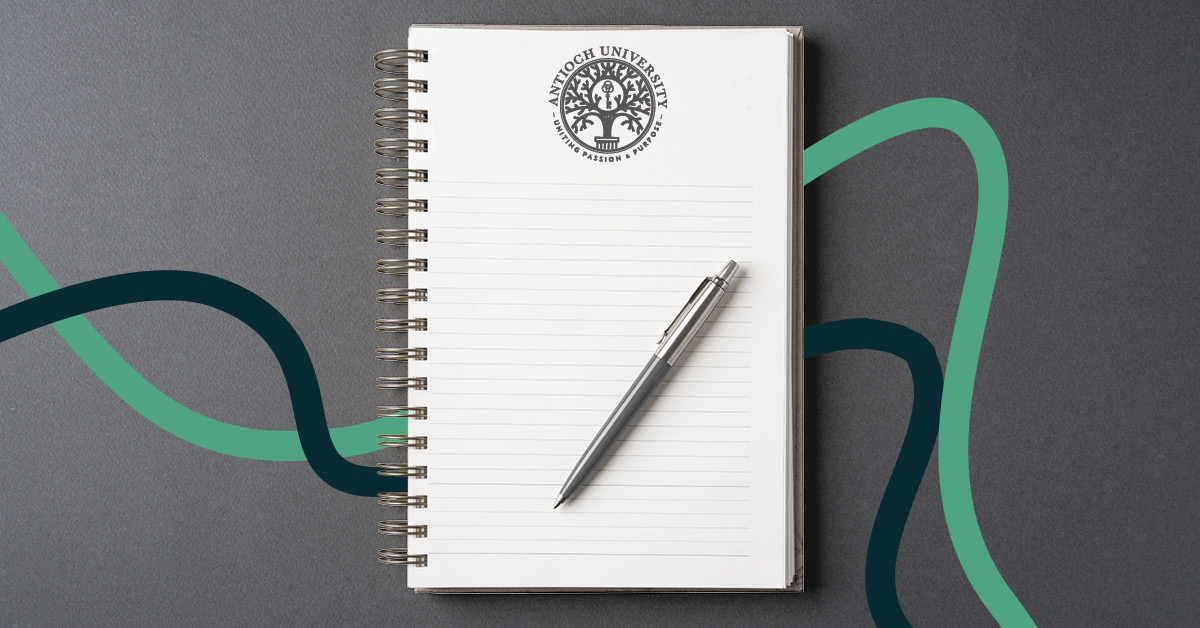 A spiral-bound notebook with lined pages lies open on a dark gray surface. The top of the page features the Antioch University logo with the words “Uniting Passion & Purpose.” A silver pen rests diagonally on the notebook, and curved teal and dark green lines flow across the background.