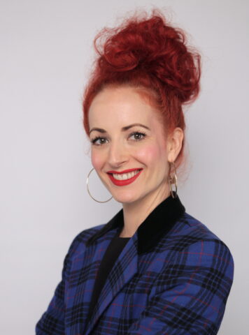Carly Petersmeyer smiling, with  red hair styled in a high curly bun, wearing hoop earrings, red lipstick, and a blue plaid jacket with a black collar, posed against a light gray background.