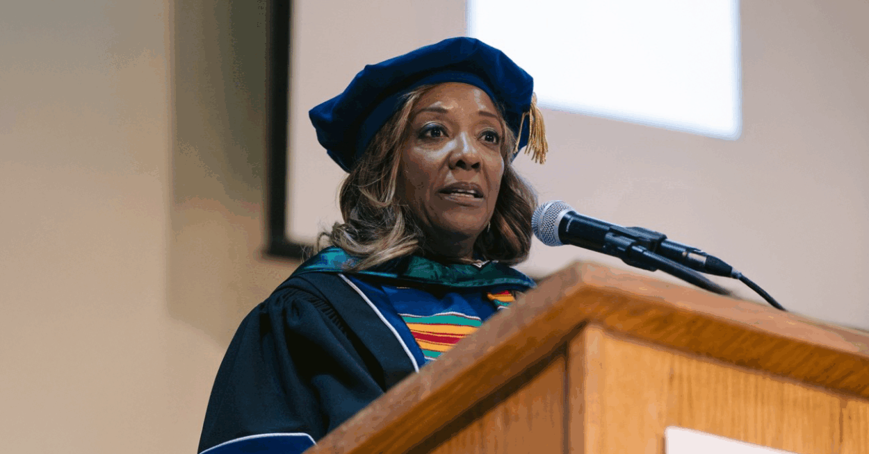 A photo of Lynne Washington speaking at a podium. She is wearing doctoral graduation regalia.