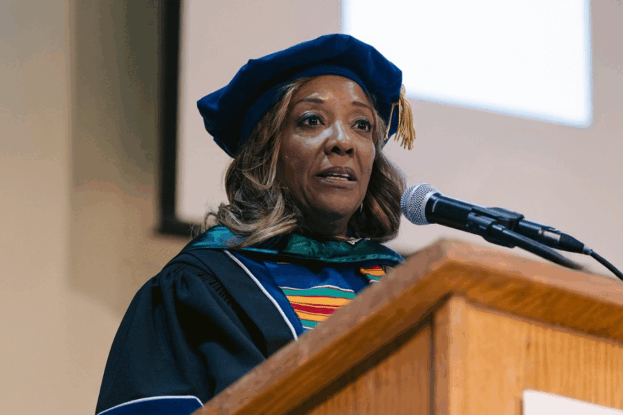 A photo of Lynne Washington speaking at a podium. She is wearing doctoral graduation regalia.