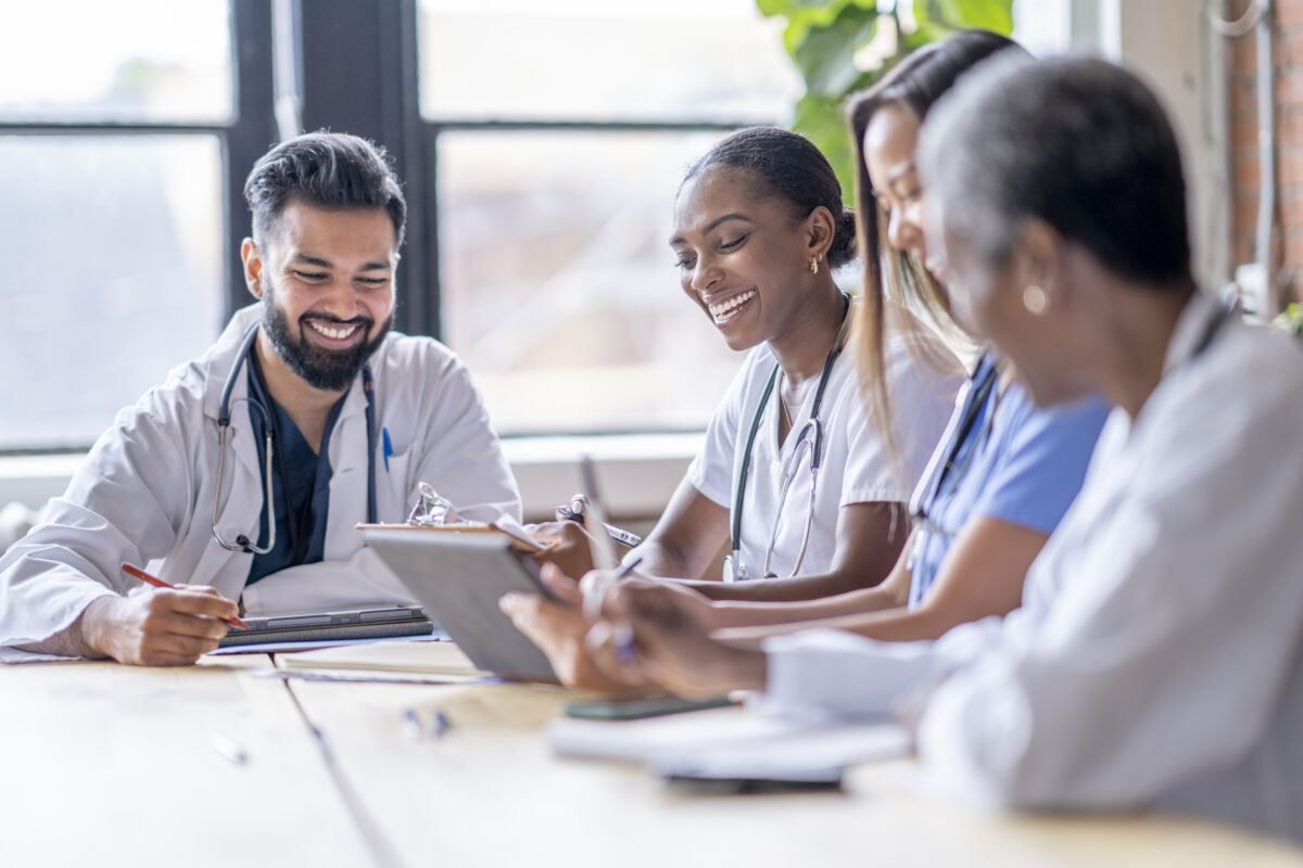 A diverse group of healthcare professionals sit together at a table, smiling and collaborating as they review notes and a digital tablet. They wear medical scrubs and lab coats with stethoscopes, creating a bright, collegial atmosphere in a well-lit room.