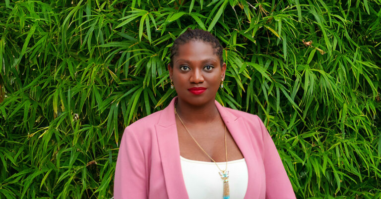 A photograph of Naydine Johney wearing a pink jacket and standing in front of leafy bamboo plants.