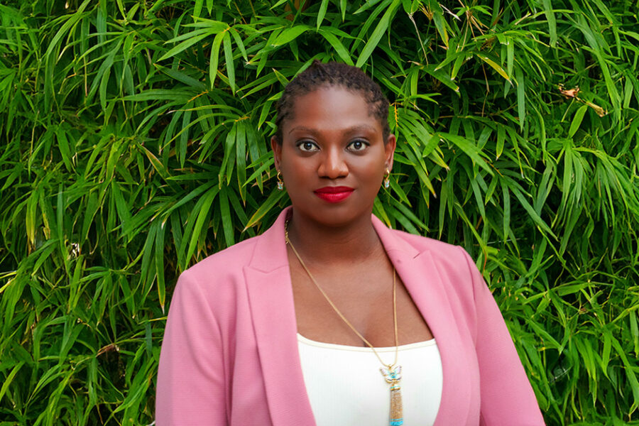 A photograph of Naydine Johney wearing a pink jacket and standing in front of leafy bamboo plants.