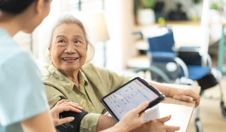 An older woman smiles warmly at a healthcare worker who is checking her blood pressure. The healthcare worker holds a tablet displaying vital sign readings while gently adjusting the blood pressure cuff on the woman's arm. A wheelchair is visible in the background of the bright, home-like setting.