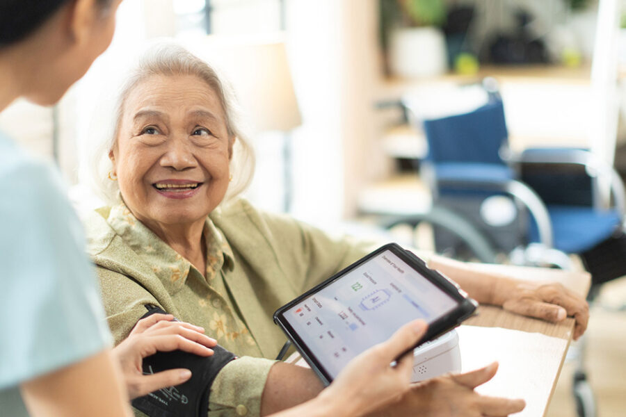 An older woman smiles warmly at a healthcare worker who is checking her blood pressure. The healthcare worker holds a tablet displaying vital sign readings while gently adjusting the blood pressure cuff on the woman's arm. A wheelchair is visible in the background of the bright, home-like setting.