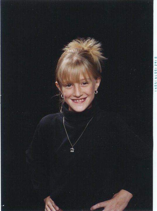 Portrait of Selina Starling as a child, smiling in a studio photo against a dark background. They have blonde hair in a messy updo with bangs and wear a black turtleneck, hoop earrings, and a necklace, with one hand resting on their hip.