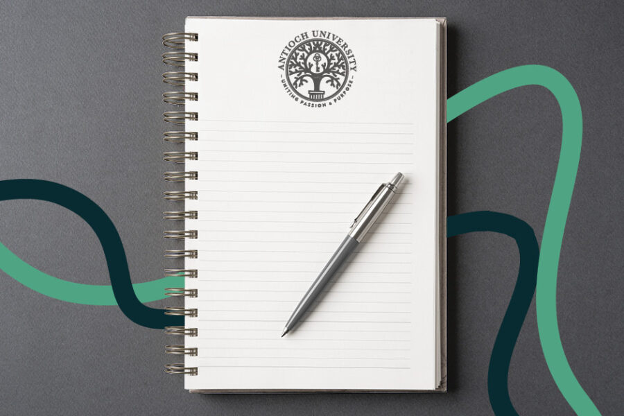 A spiral-bound, lined notebook with the Antioch University seal on the cover sits on a gray surface with a silver pen across it, framed by teal and dark green wavy lines.