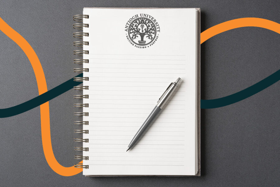 Alt text: A spiral-bound notebook with the Antioch University logo sits on a dark gray surface with a silver pen on the page. Curving teal and orange lines flow behind the notebook across the background.