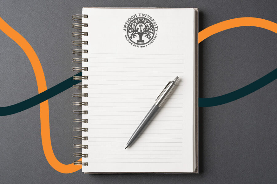 Spiral-bound notebook with the Antioch University seal on lined pages and a pen resting on top, set against a gray background with flowing orange and dark teal lines