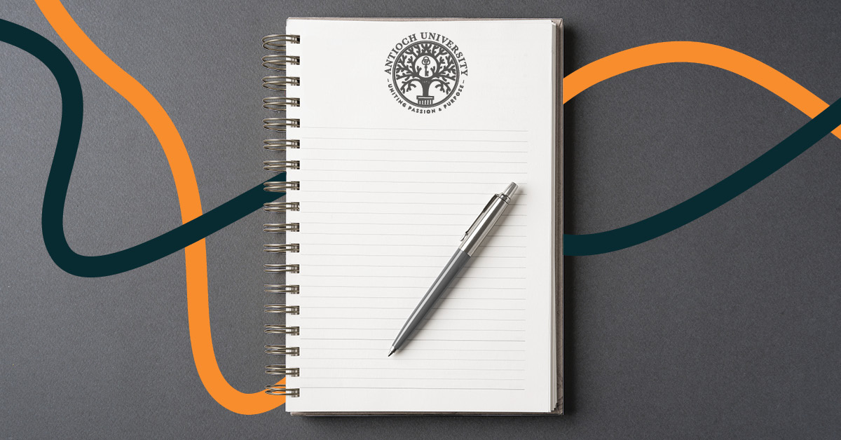 Spiral-bound notebook with the Antioch University seal on lined pages and a pen resting on top, set against a gray background with flowing orange and dark teal lines