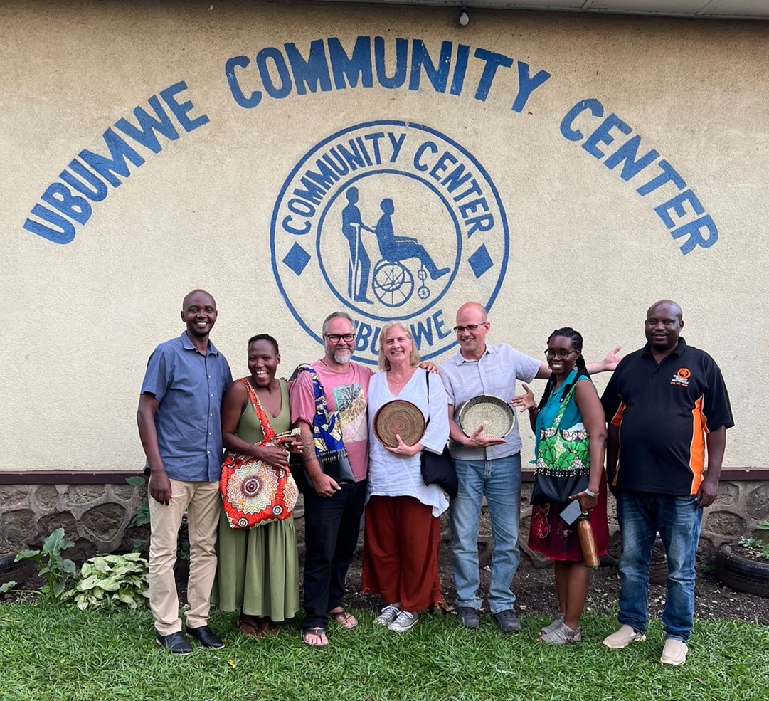 Group of eight people standing outdoors in front of a wall painted with ‘Ubumwe Community Center’ and a circular logo, smiling and posing together; several hold woven baskets, with grass and plants in the foreground