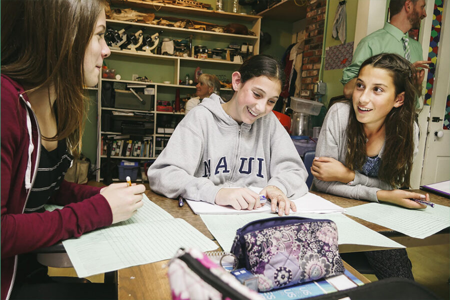Three students sit around a table working together on papers, smiling and talking in a classroom while a teacher stands in the background.