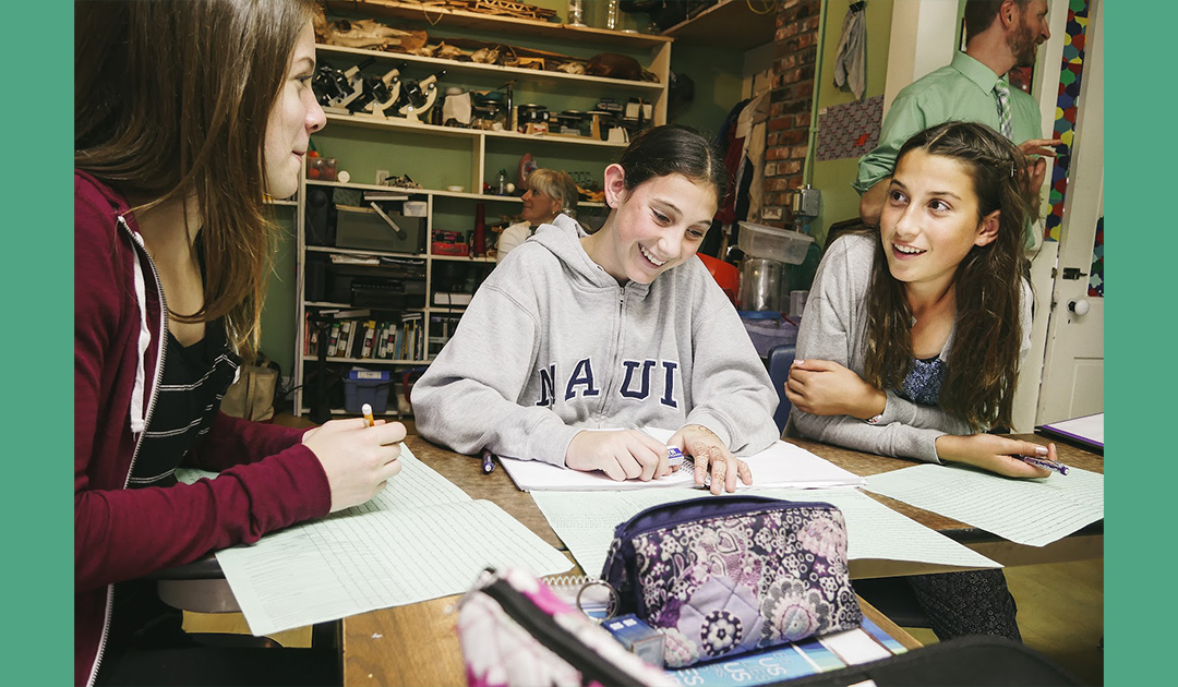 Three students sit around a table working together on papers, smiling and talking in a classroom while a teacher stands in the background.