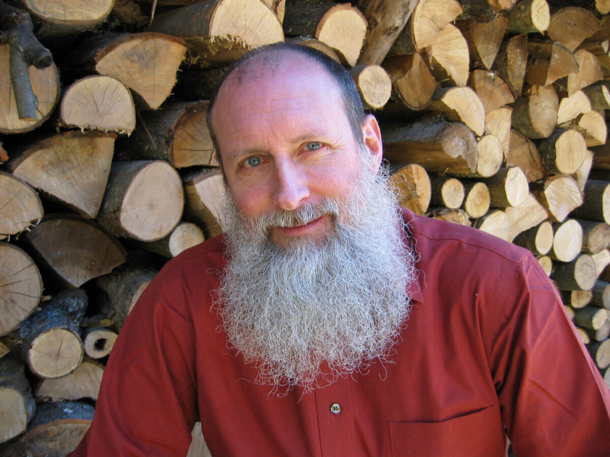 Tom Wessels, environmental educator and author, smiling in front of a stacked woodpile, wearing a red button-down shirt.
