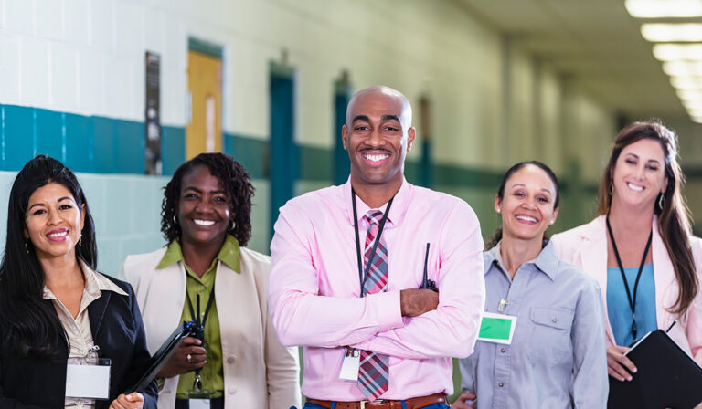 A multiracial group of five teachers or school administrators standing together in a school hallway smiling confidently at the camera in a group portrait. The African-American man in the middle is the team leader, perhaps the school principal. He is the only male in the group. The focus is on him and the Hispanic woman standing on the left. Representative of Inclusive Learning