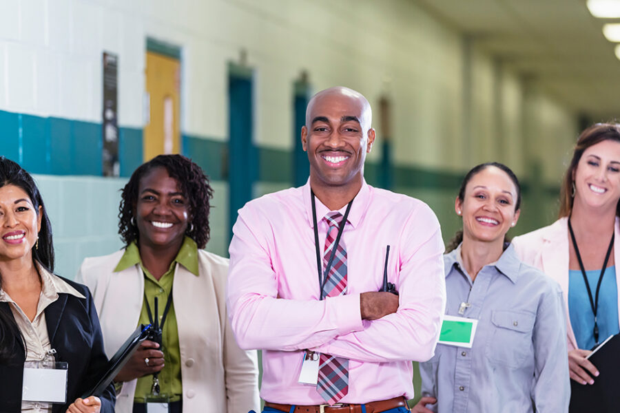 A multiracial group of five teachers or school administrators standing together in a school hallway smiling confidently at the camera in a group portrait. The African-American man in the middle is the team leader, perhaps the school principal. He is the only male in the group. The focus is on him and the Hispanic woman standing on the left. Representative of Inclusive Learning