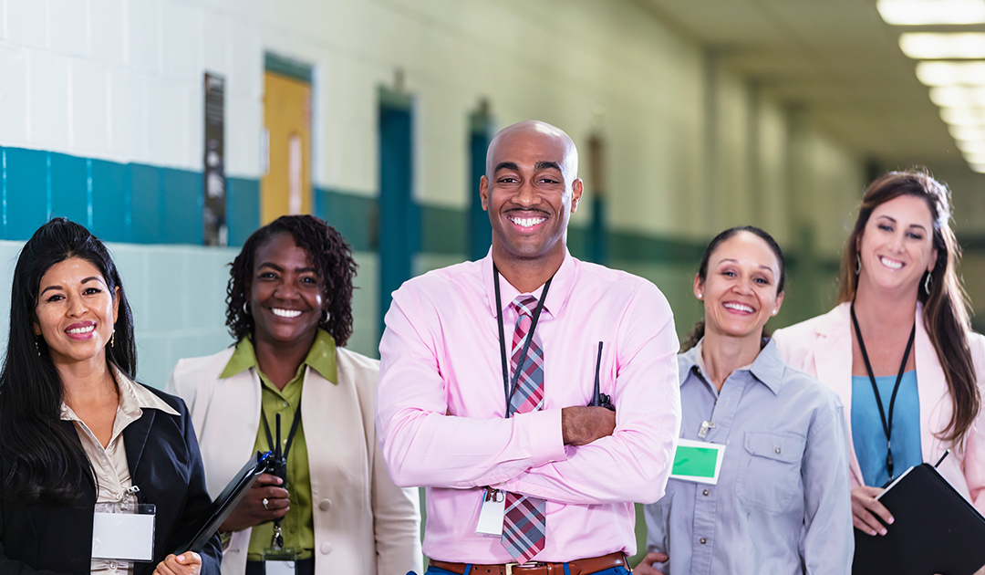 A multiracial group of five teachers or school administrators standing together in a school hallway smiling confidently at the camera in a group portrait. The African-American man in the middle is the team leader, perhaps the school principal. He is the only male in the group. The focus is on him and the Hispanic woman standing on the left. Representative of Inclusive Learning