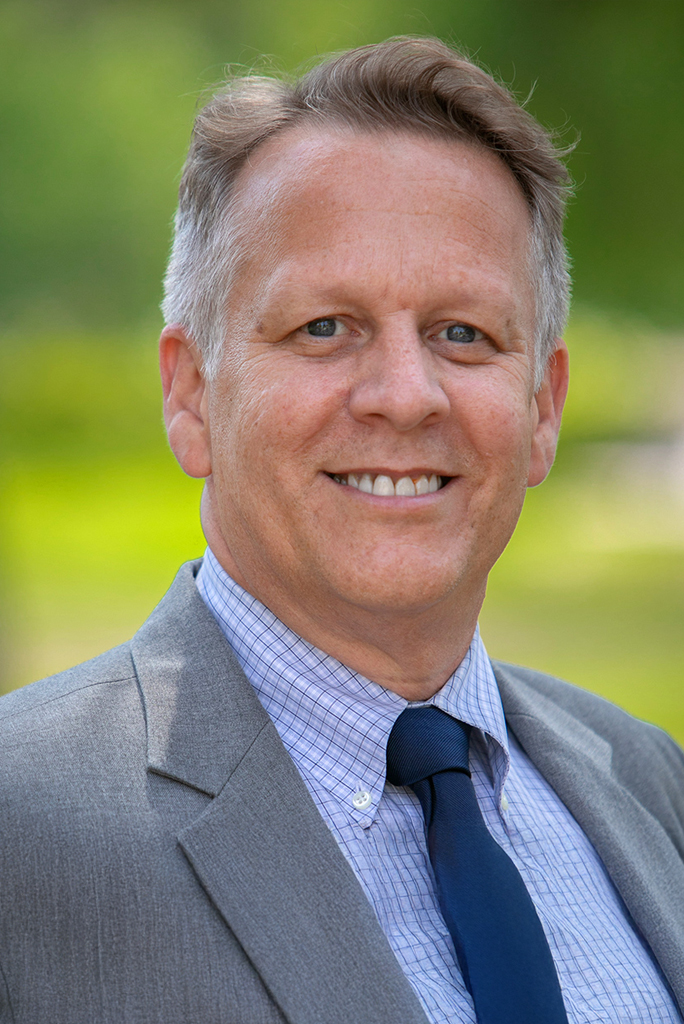 A professional headshot of Tom Hoener smiling at the camera. He has short light brown hair and is wearing a gray suit jacket, a light blue checkered dress shirt, and a dark blue tie, with a blurred green outdoor background.