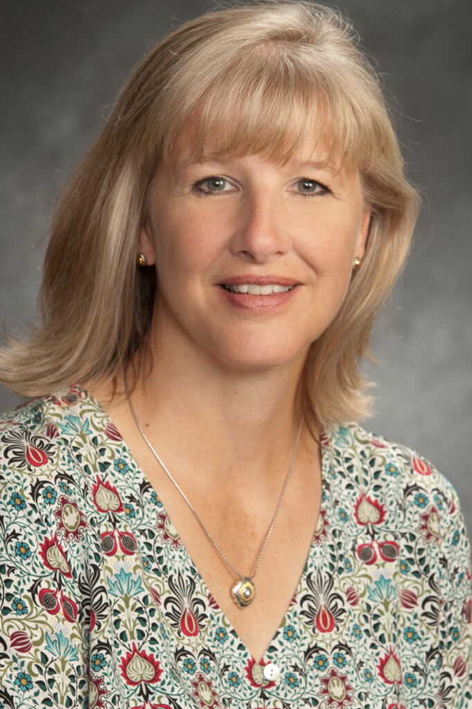 Sandra Madar, a woman with shoulder-length blonde hair and bangs, smiling at the camera in a professional headshot. She is wearing a patterned blouse with red, blue, and green details, small gold earrings, and a necklace, against a neutral gray studio background.
