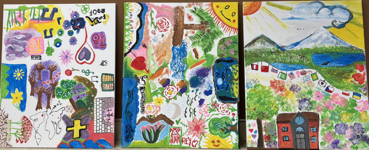 Three colorful, abstract paintings displayed side by side, featuring a mix of symbols, landscapes, flowers, faces, a sun, mountains, and a building with international flags, created in a vibrant, collaborative style.