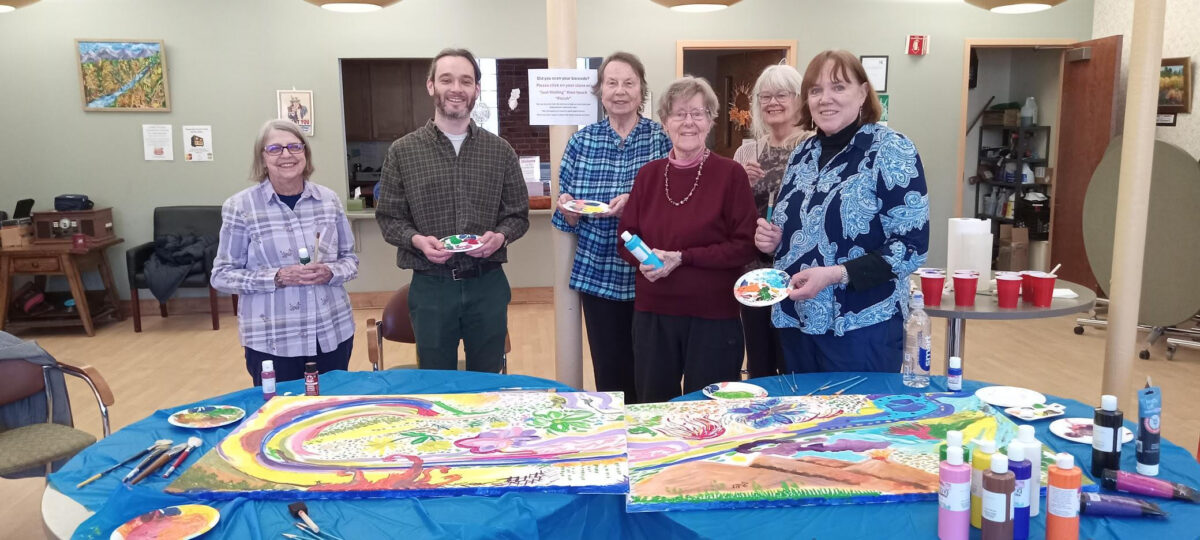 Group of seven adults standing around a table at a community center, each holding paint or small palettes, smiling behind a large, colorful collaborative painting spread across the table with brushes, paint bottles, and supplies visible.