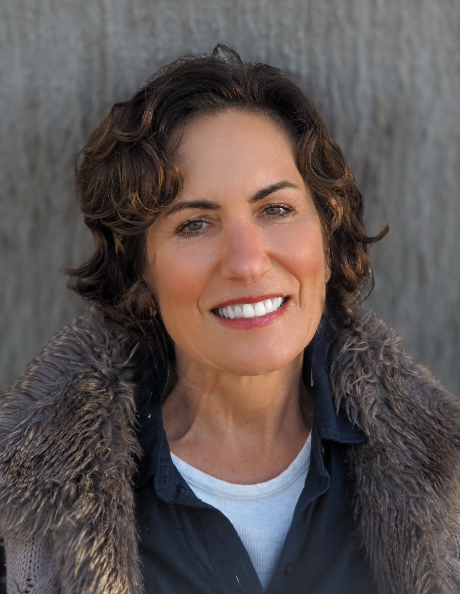 Professional headshot of Lisa Richter smiling warmly, with short curly dark hair, wearing a dark collared shirt and a textured jacket, photographed outdoors with a soft, blurred background.