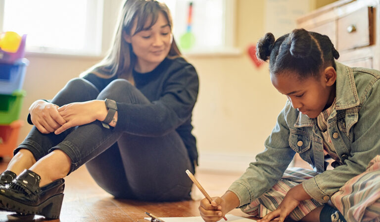 An art therapist working with a girl drawing on paper on the floor.