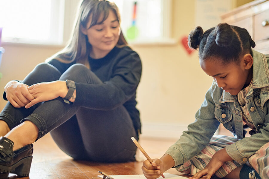 An art therapist working with a girl drawing on paper on the floor.