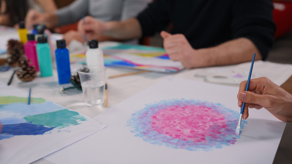 People working at a table with art supplies. Their hands and bodies are visible. One painter is painting a blue and pink circle with dots.