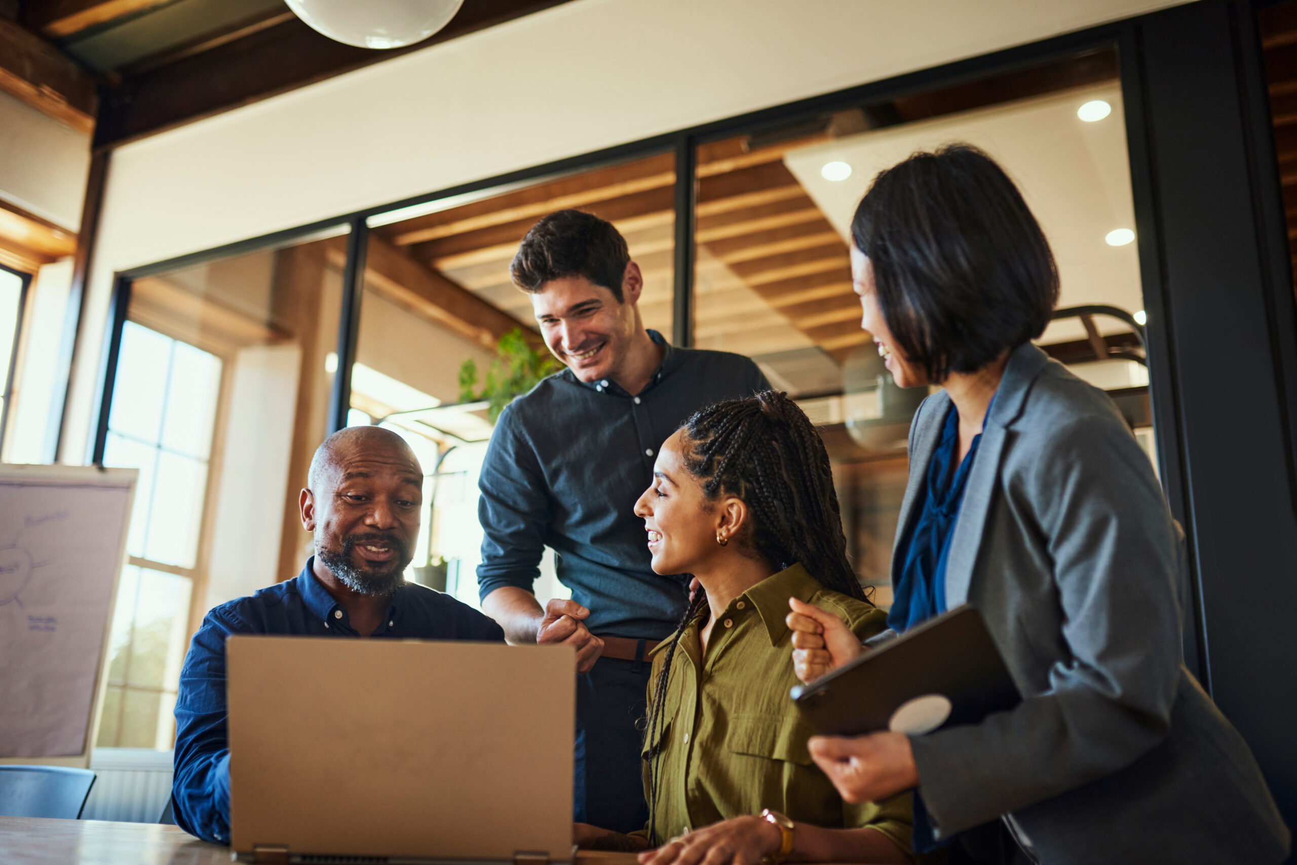 A diverse group of professionals gathered around a laptop, smiling and collaborating in a modern office setting.