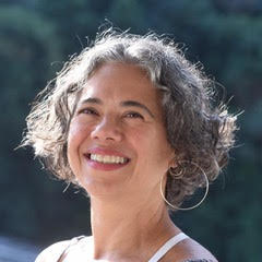 Portrait of Giselle Ruzany smiling warmly outdoors, with short, curly gray hair and large hoop earrings. She is wearing a light-colored top and looking slightly upward, with soft natural light illuminating her face against a blurred green background.