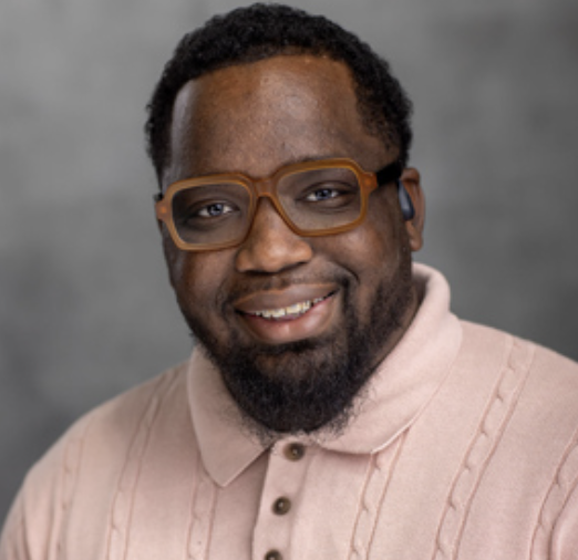 Portrait of Bernell L. Elzey Jr., wearing brown-framed glasses and a light pink collared sweater against neutral gray background.