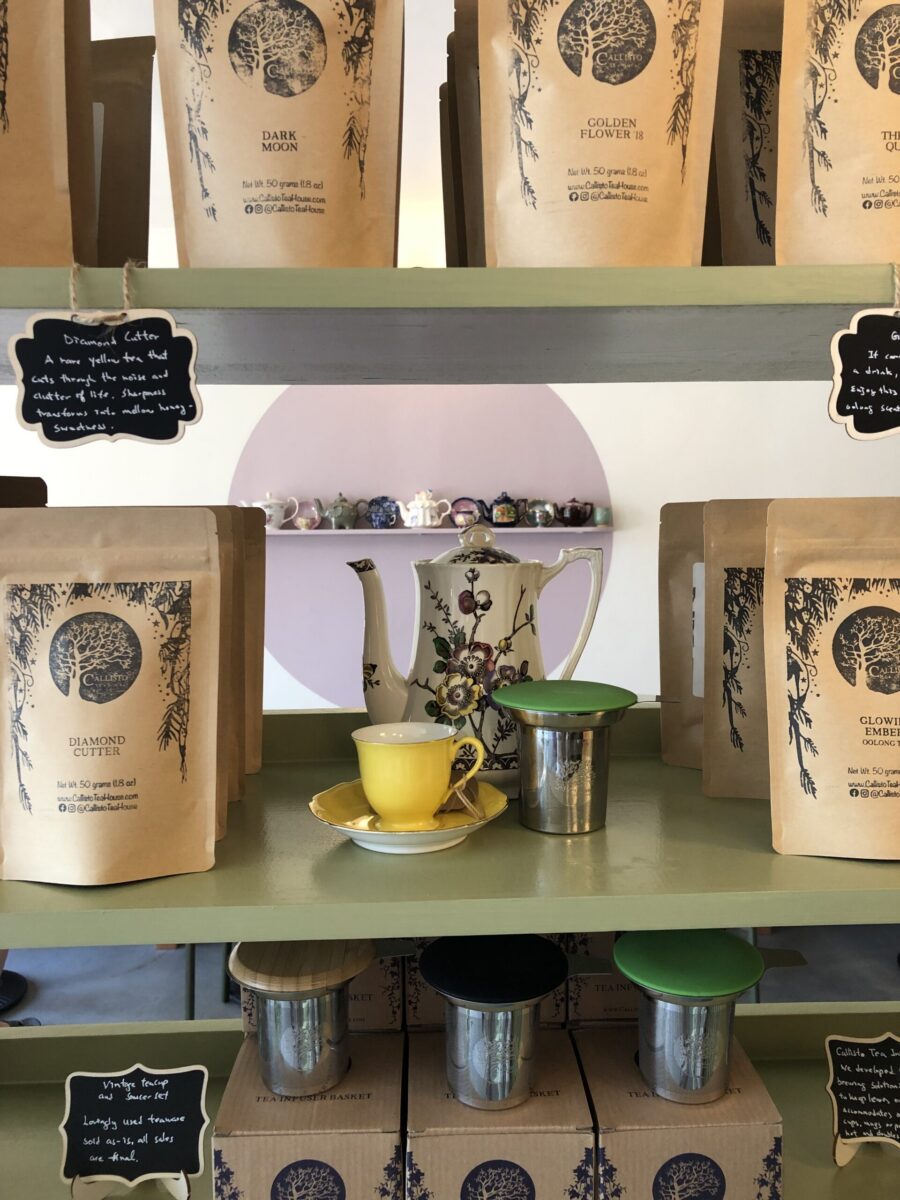 Shelves inside Callisto Tea House displaying packaged loose-leaf teas, vintage teapots, tea infusers, and a bright yellow teacup and saucer arranged around a floral teapot. Handwritten signs describing tea blends and tea accessories add to the warm, curated atmosphere of the Pasadena tea shop.