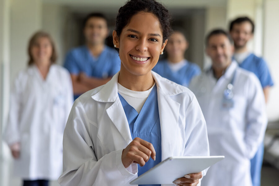 A smiling healthcare professional in a white coat and scrubs holds a tablet while standing in a hospital hallway with a medical team in the background.