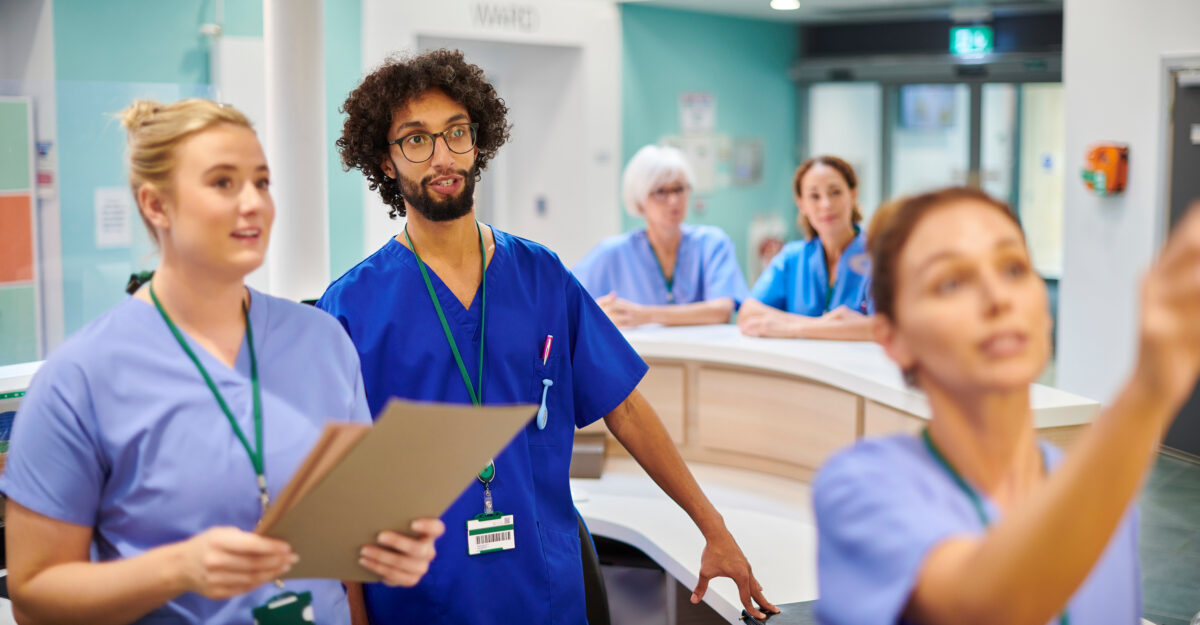 A diverse group of healthcare professionals in scrubs collaborates at a hospital nurses’ station, reviewing patient information during a team discussion.