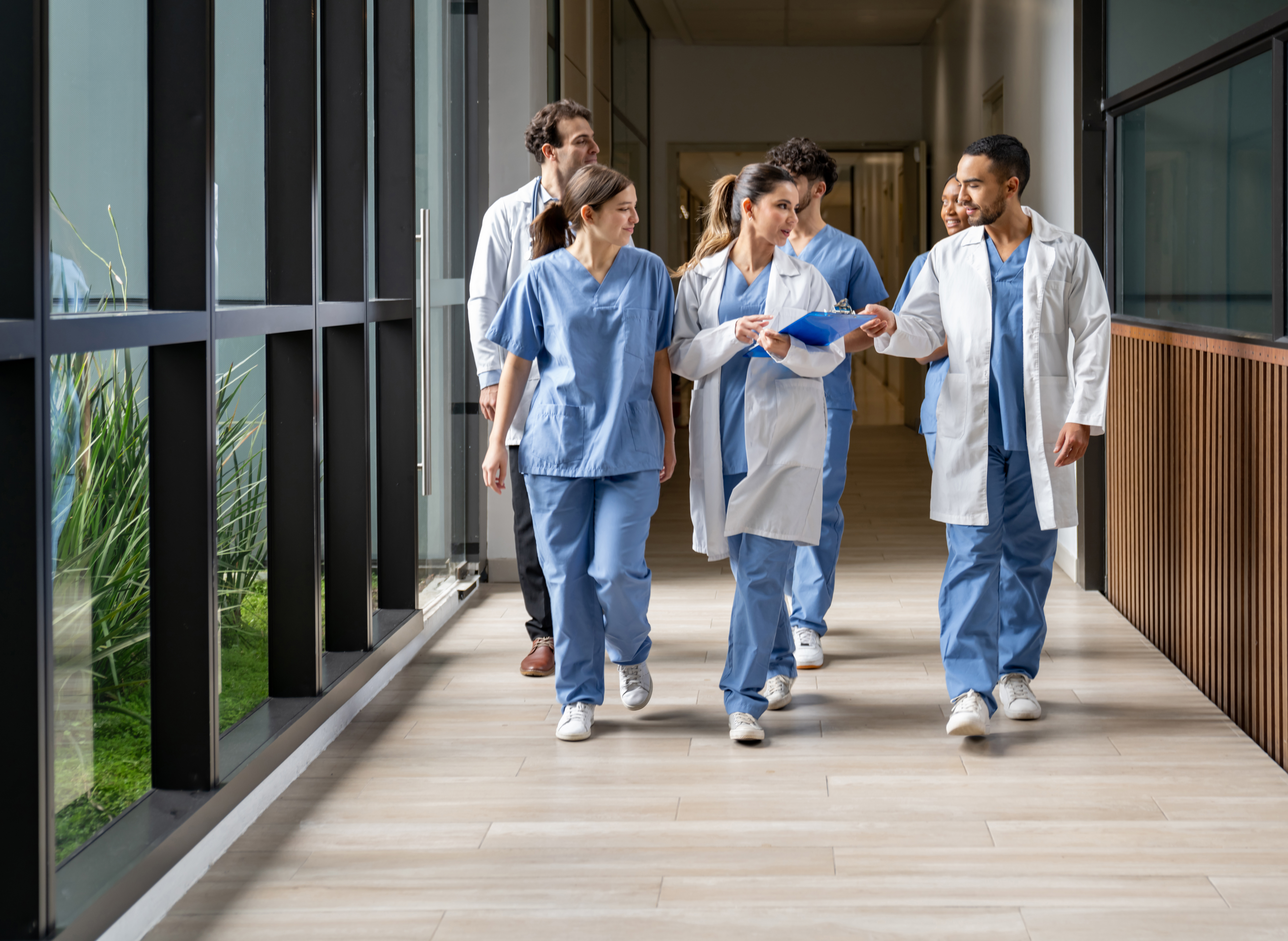 A group of healthcare professionals in scrubs and lab coats walk down a bright hospital hallway, collaborating and reviewing information on a clipboard.