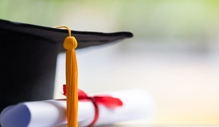 Close-up of a graduation cap with a gold tassel and rolled diploma tied with a red ribbon against a soft blurred background.