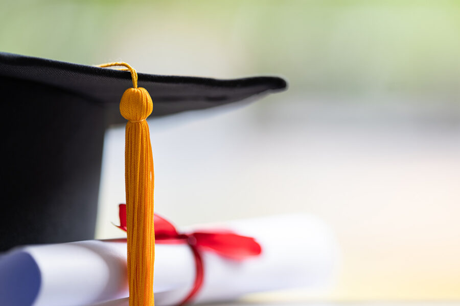 Close-up of a graduation cap with a gold tassel and rolled diploma tied with a red ribbon against a soft blurred background.