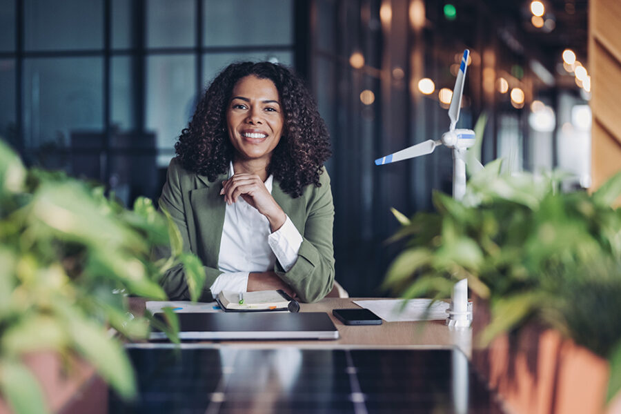 Smiling professional seated in a modern office surrounded by plants with wind turbine models and solar panels, representing sustainable leadership and clean energy innovation.