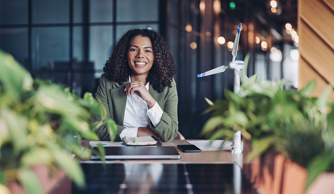 Smiling professional seated in a modern office surrounded by plants with wind turbine models and solar panels, representing sustainable leadership and clean energy innovation.