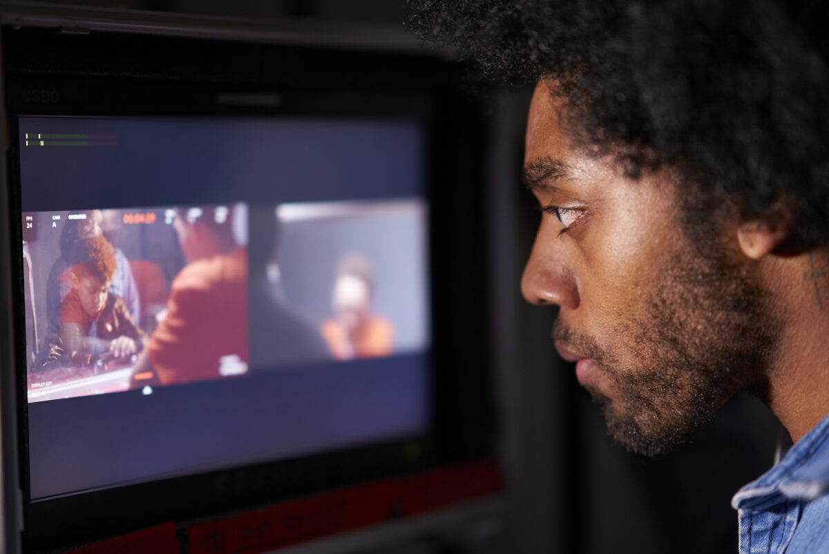 Close-up profile of a person watching a monitor displaying a group of people filming a scene in a dim room.