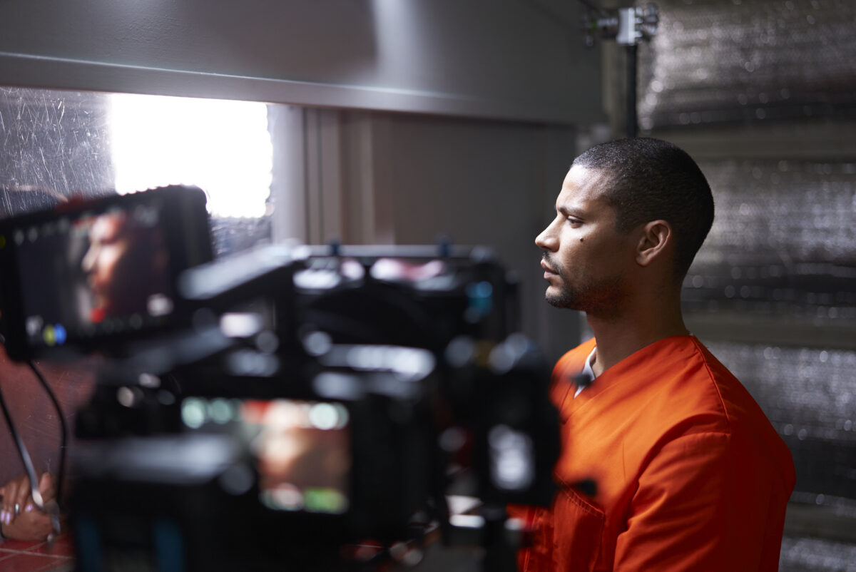 A man wearing an orange shirt sits in profile on a film set while several cameras and monitors record him.