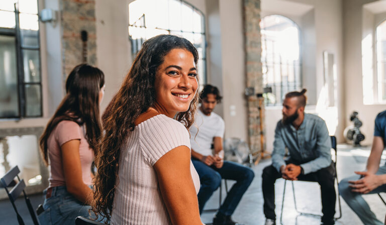 Portrait of a woman looking at camera during a group therapy session. Mental health concept in a loft with big windows. Healthcare and medicine concept. Group of people sitting in a circle are participating in a support meeting.