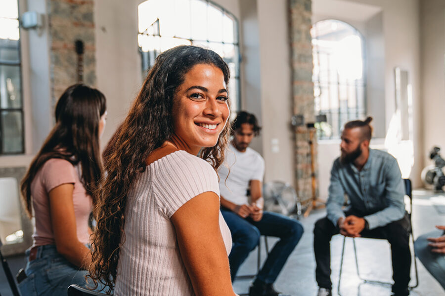 Portrait of a woman looking at camera during a group therapy session. Mental health concept in a loft with big windows. Healthcare and medicine concept. Group of people sitting in a circle are participating in a support meeting.