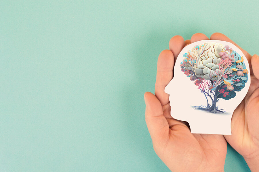 Two hands gently cradle a stylized silhouette of a human head against a soft teal background. Inside the head is an artistic illustration of a brain intertwined with colorful tree branches and foliage, symbolizing mental health, personal growth, healing, and the connection between mind and nature.