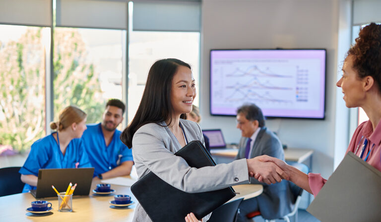 Healthcare administrator shaking hands with a colleague during a meeting, with medical staff collaborating in the background.