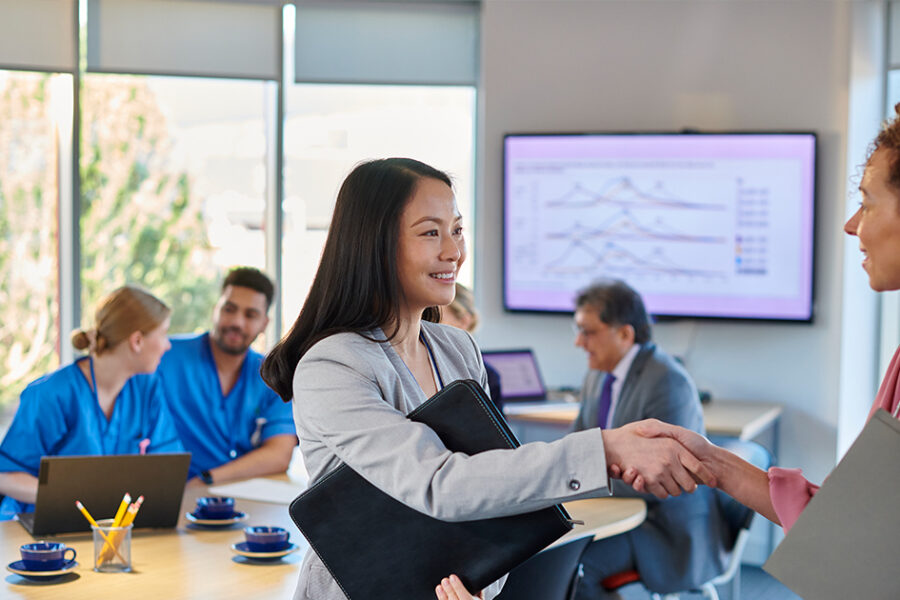 Healthcare administrator shaking hands with a colleague during a meeting, with medical staff collaborating in the background.