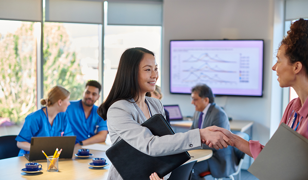 Healthcare administrator shaking hands with a colleague during a meeting, with medical staff collaborating in the background.