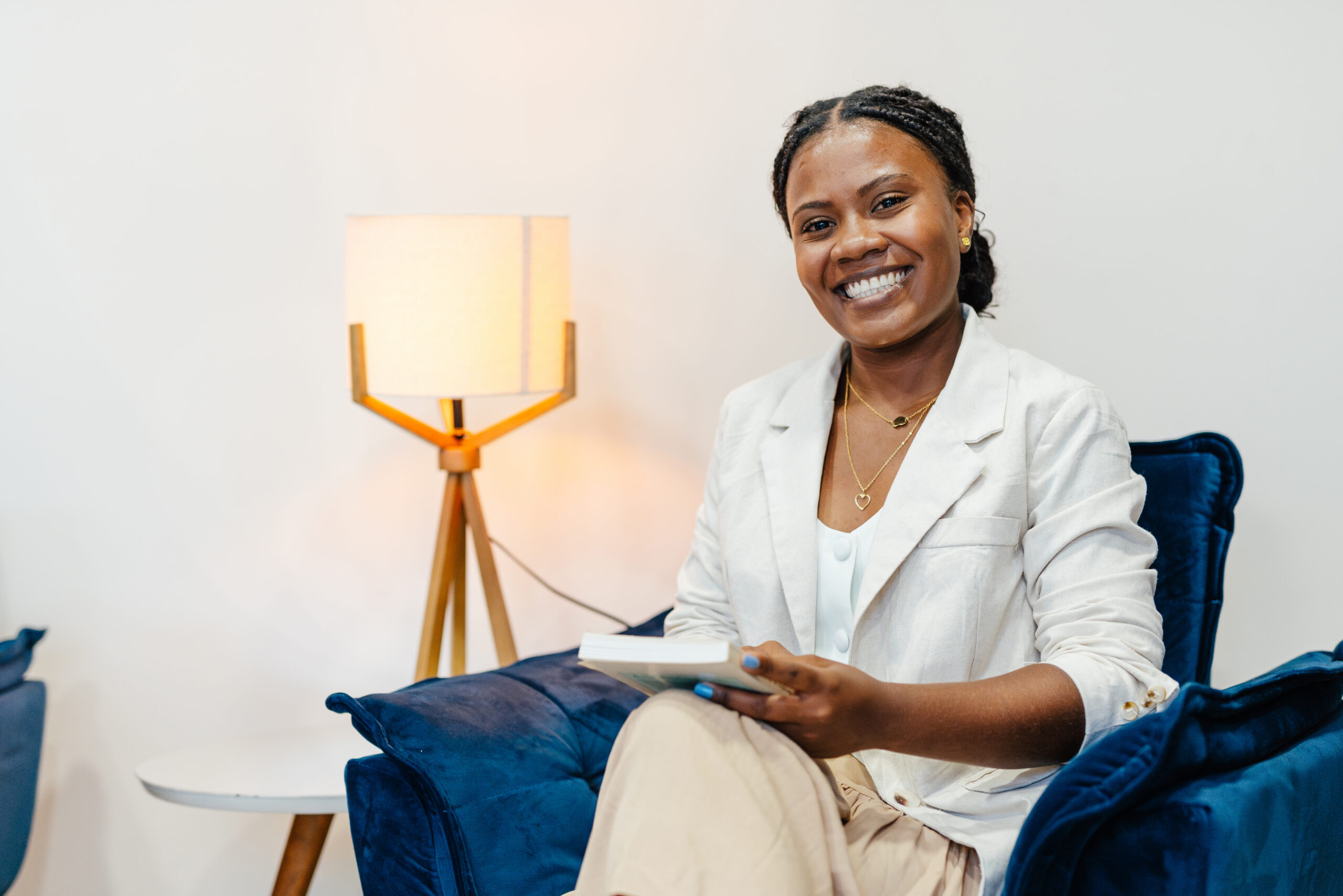 Smiling woman in a light blazer seated on a blue chair, holding a notebook with a lamp in the background.